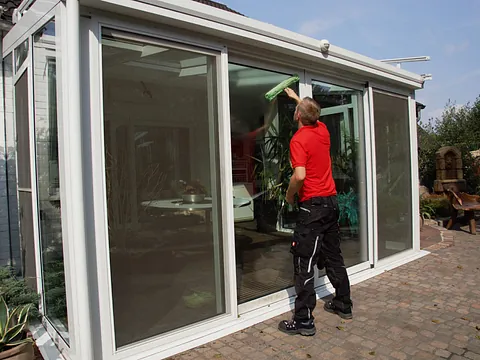 Mann in rotem Shirt reinigt die Glasfront eines Wintergartens mit einem grünen Tuch