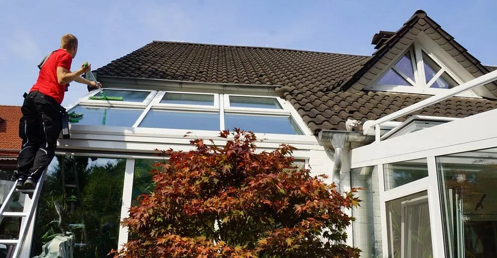Mann in rotem T-Shirt reinigt Fenster eines Wintergartens an einem Haus mit braunem Ziegeldach
