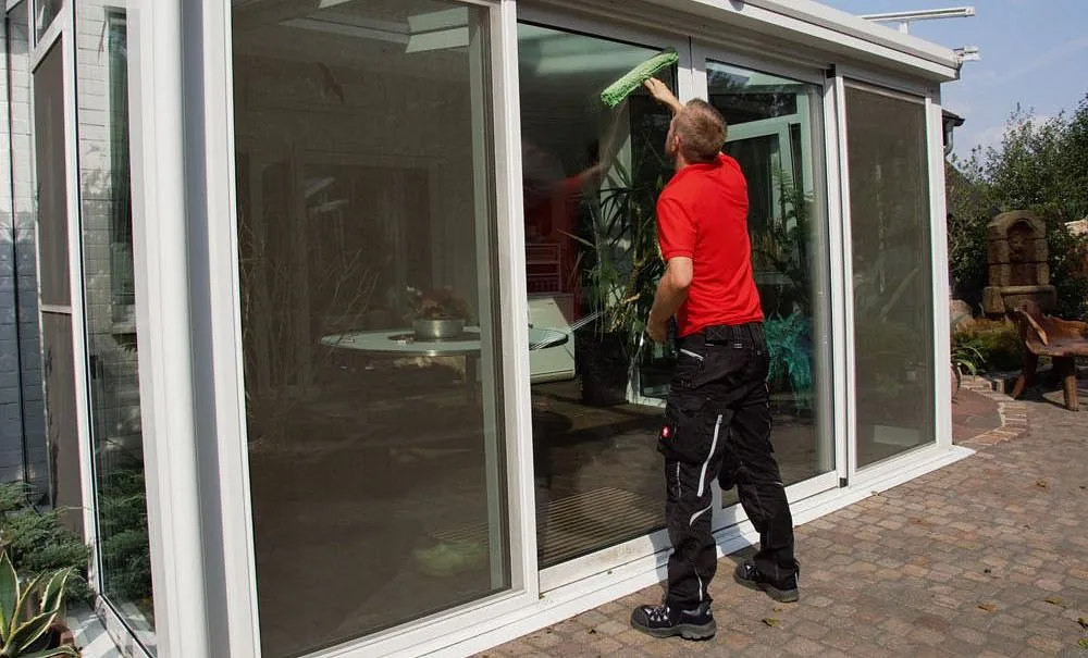 Mann in rotem T-Shirt reinigt große Glasfenster eines Wintergartens mit einem Fensterwischer