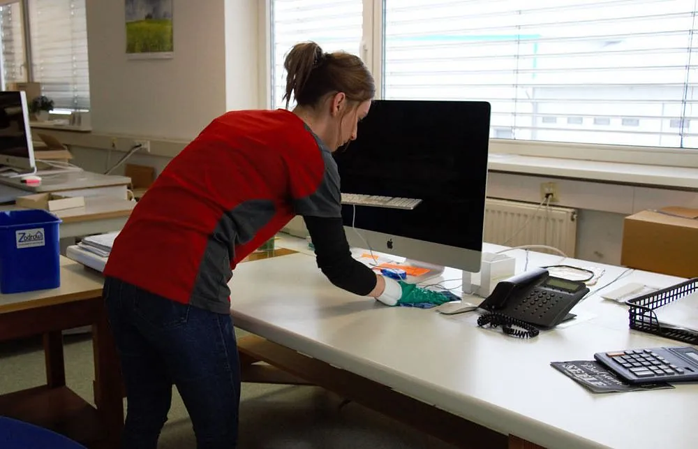 Person wischt mit Tuch einen weißen Schreibtisch mit Computer, Telefon und Tastatur in einem Büro
