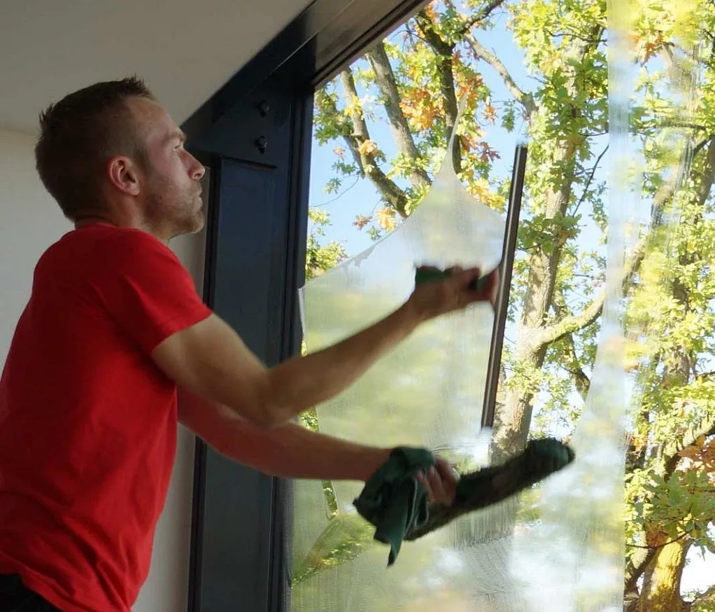Mann in rotem T-Shirt reinigt großes Fenster mit Abzieher und Tuch, draußen Bäume mit Herbstlaub sichtbar